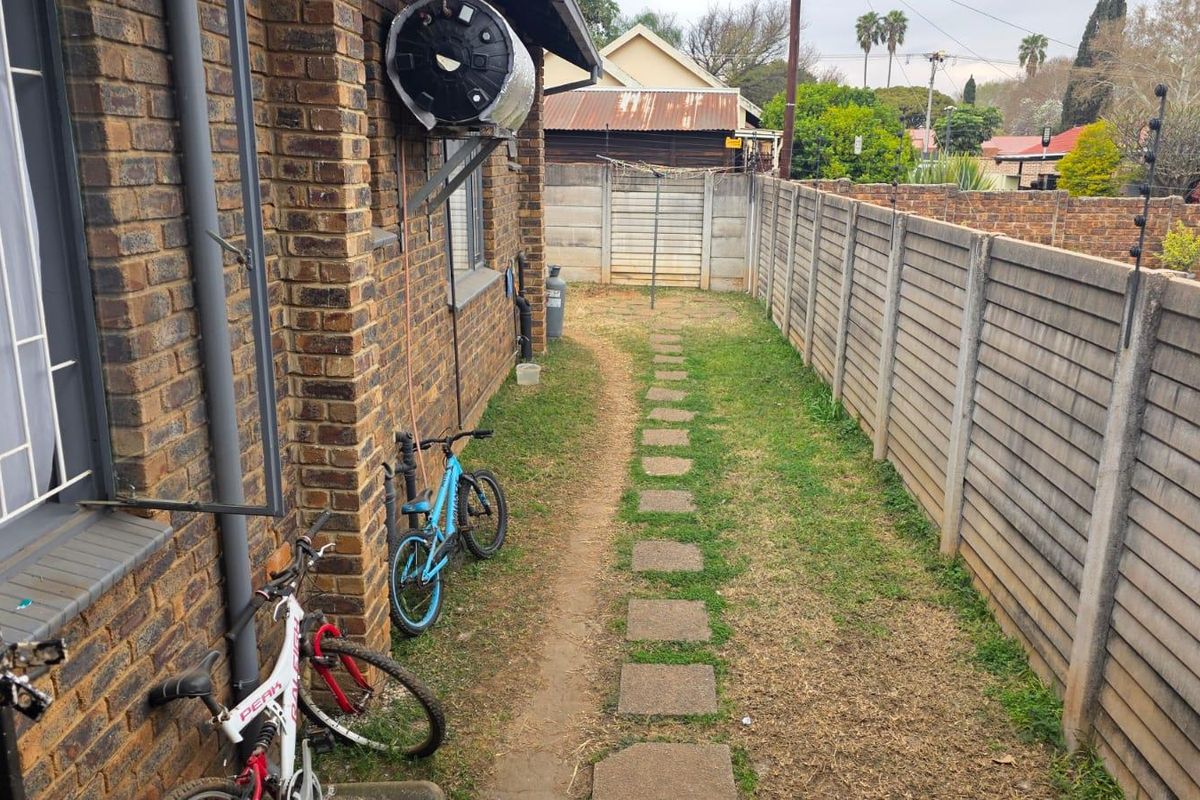 A narrow rear garden area featuring a brick exterior wall, solar geyser, precast concrete boundary fencing, and a stepping stone pathway.