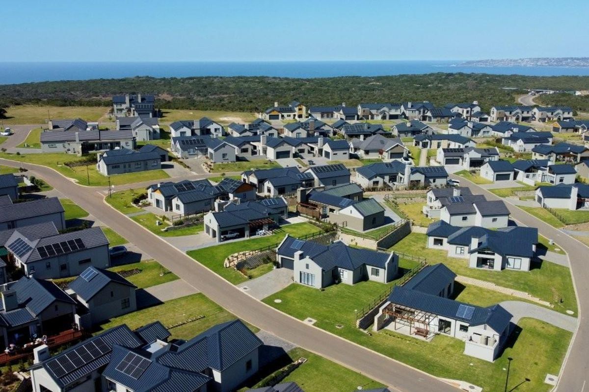 An aerial view of a modern residential estate featuring contemporary homes with dark tiled roofs, paved streets, and landscaped gardens near the coastline.
