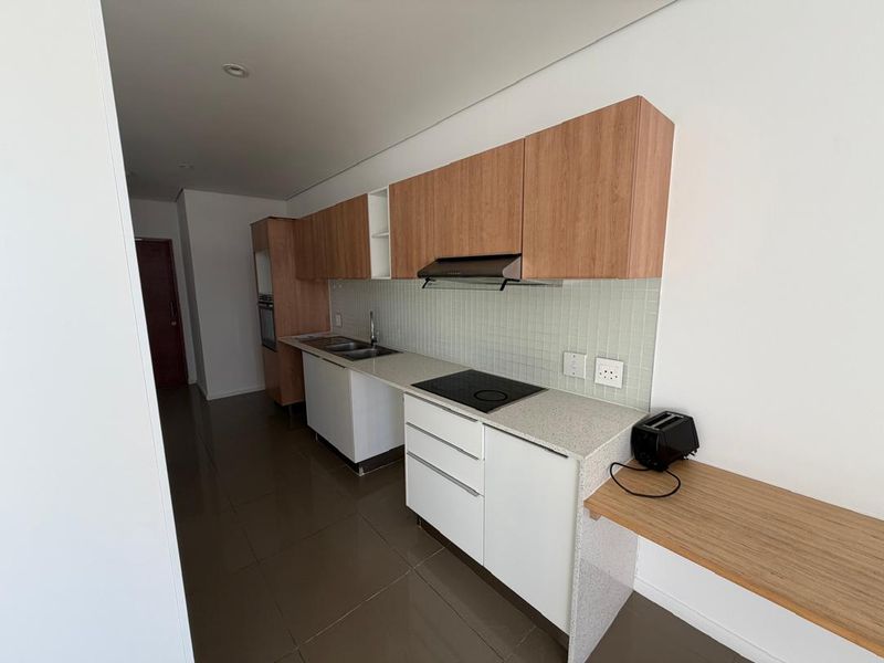 Modern galley-style kitchen featuring light wood-tone cabinetry, stone countertops, integrated electric hob, extractor fan, and sleek tiled backsplash.