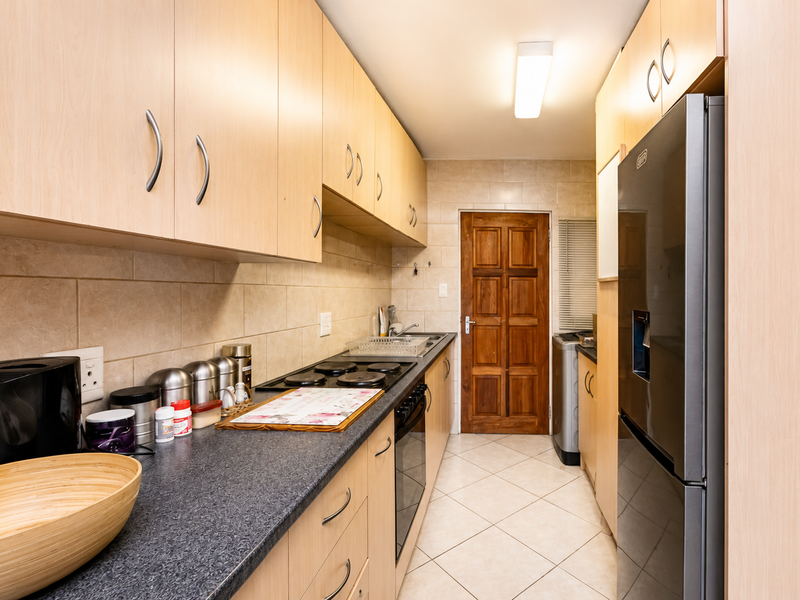 A functional galley-style kitchen featuring light wood cabinetry, dark laminate countertops, tiled backsplash, and built-in electric cooking appliances.