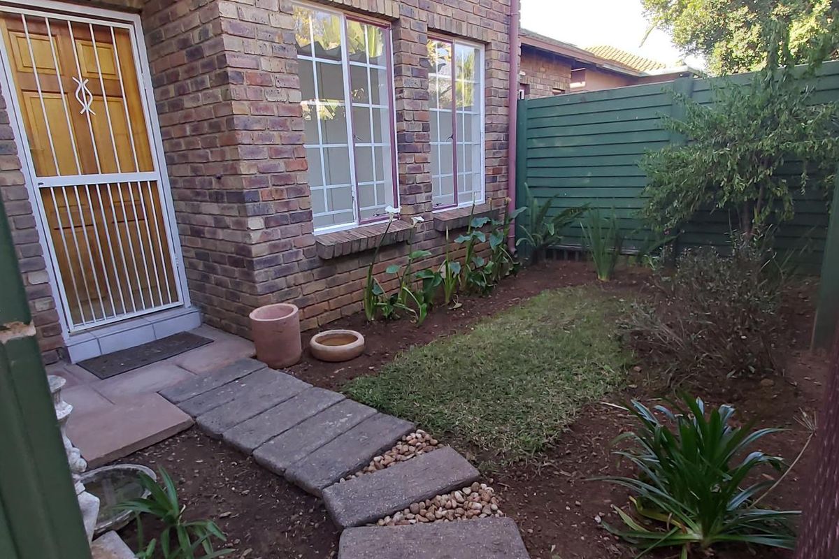A rear exterior view of a face brick townhouse featuring a small garden area, paved walkway, and secure gated access.