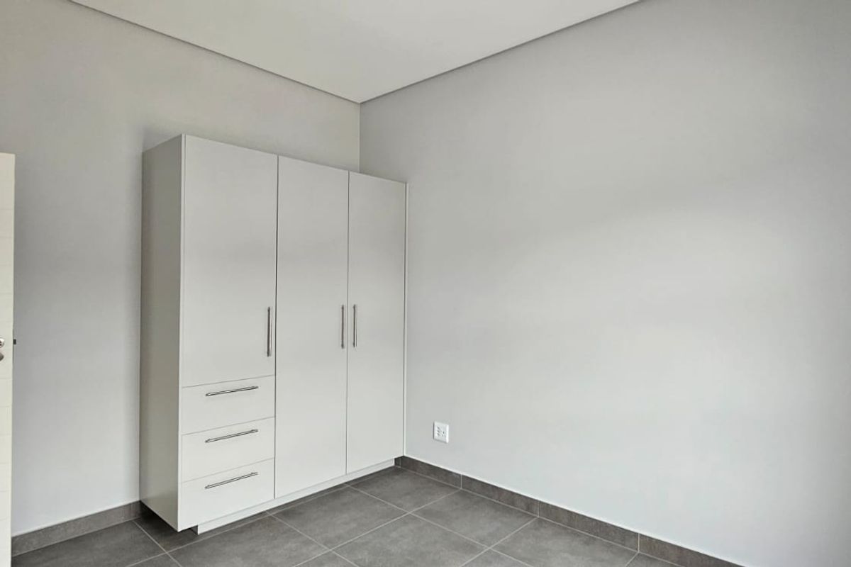A clean, modern bedroom featuring neutral grey floor tiles, crisp white walls, and a functional built-in wardrobe with integrated storage.