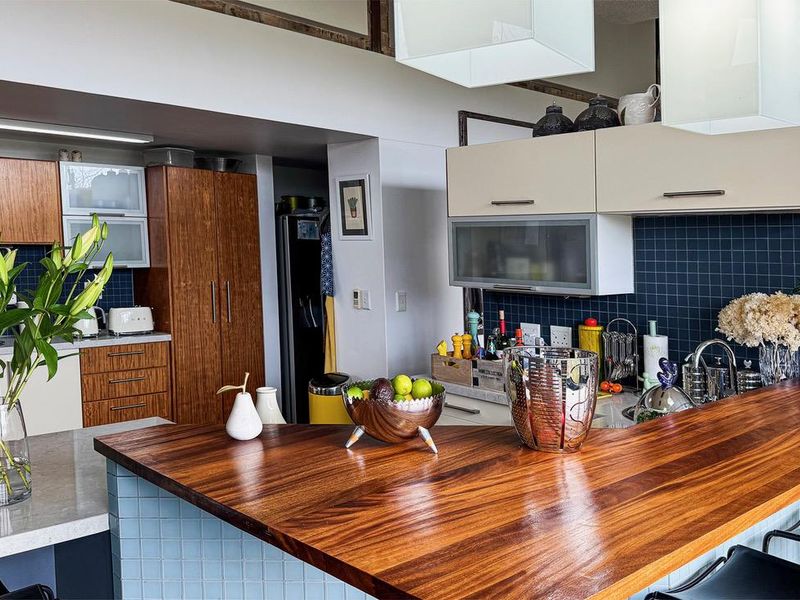 A modern kitchen featuring a large wooden breakfast bar, stone countertops, dark tiled backsplash, and ample built-in cabinetry with integrated lighting.