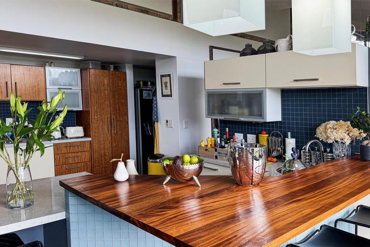 A modern kitchen featuring a large wooden breakfast bar, stone countertops, dark tiled backsplash, and ample built-in cabinetry with integrated lighting.