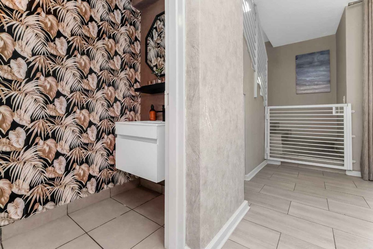 A compact guest bathroom featuring a wall-mounted vanity, patterned wallpaper, and tiled flooring, adjacent to a hallway with a safety gate.