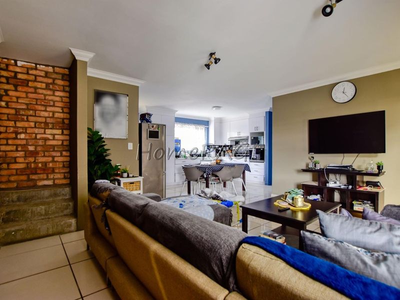 Open-plan living and kitchen area featuring tiled floors, an exposed brick feature wall, and white cabinetry in the kitchen space.