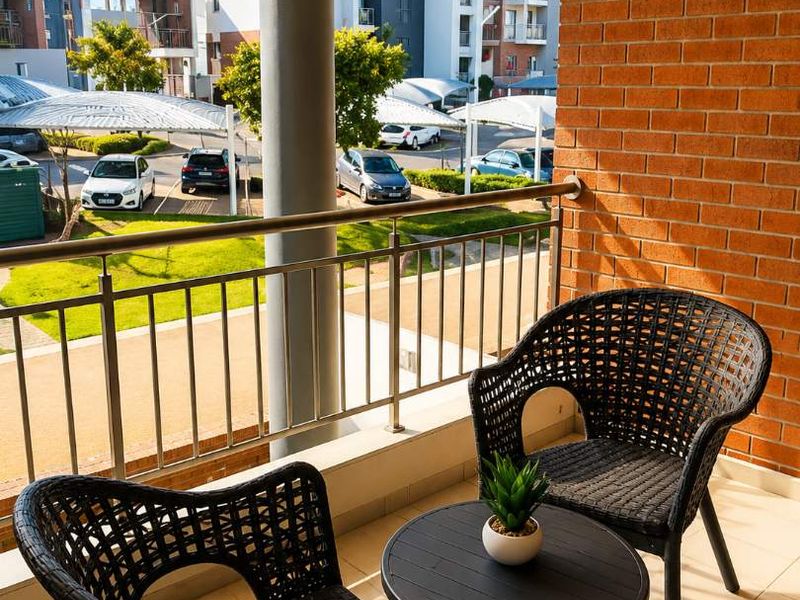 A covered balcony featuring a brick accent wall, tiled flooring, and a metal balustrade overlooking the apartment complex grounds.