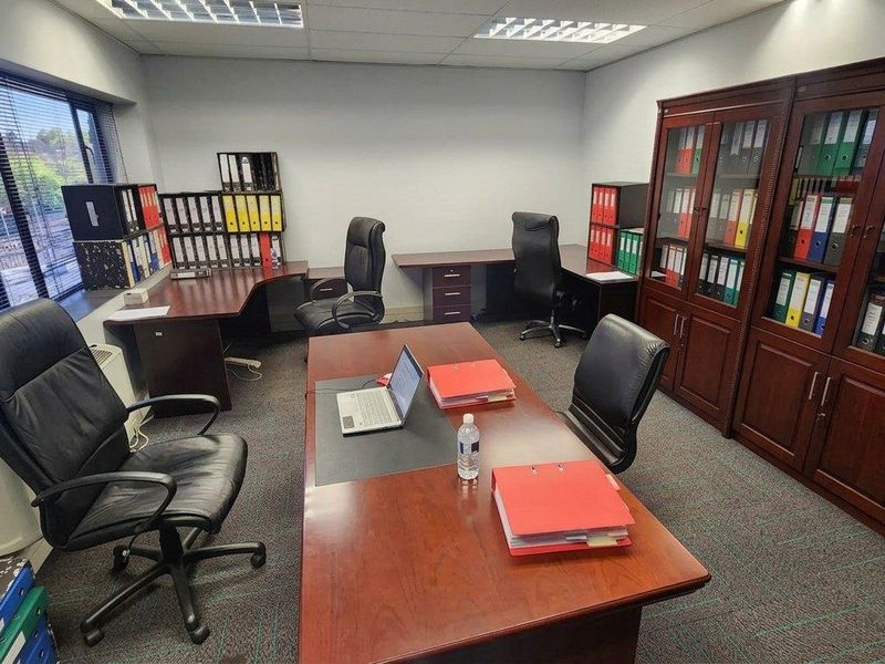 A professional office space featuring built-in wooden storage cabinetry, carpeted flooring, and large windows with venetian blinds, providing a functional workspace environment.