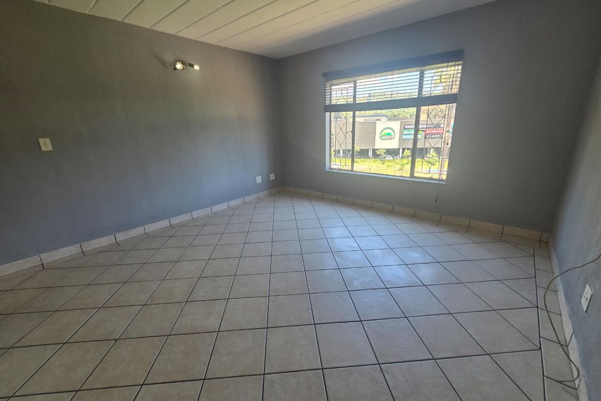 An empty living room featuring neutral grey walls, tiled flooring, and a large window with security bars and venetian blinds.