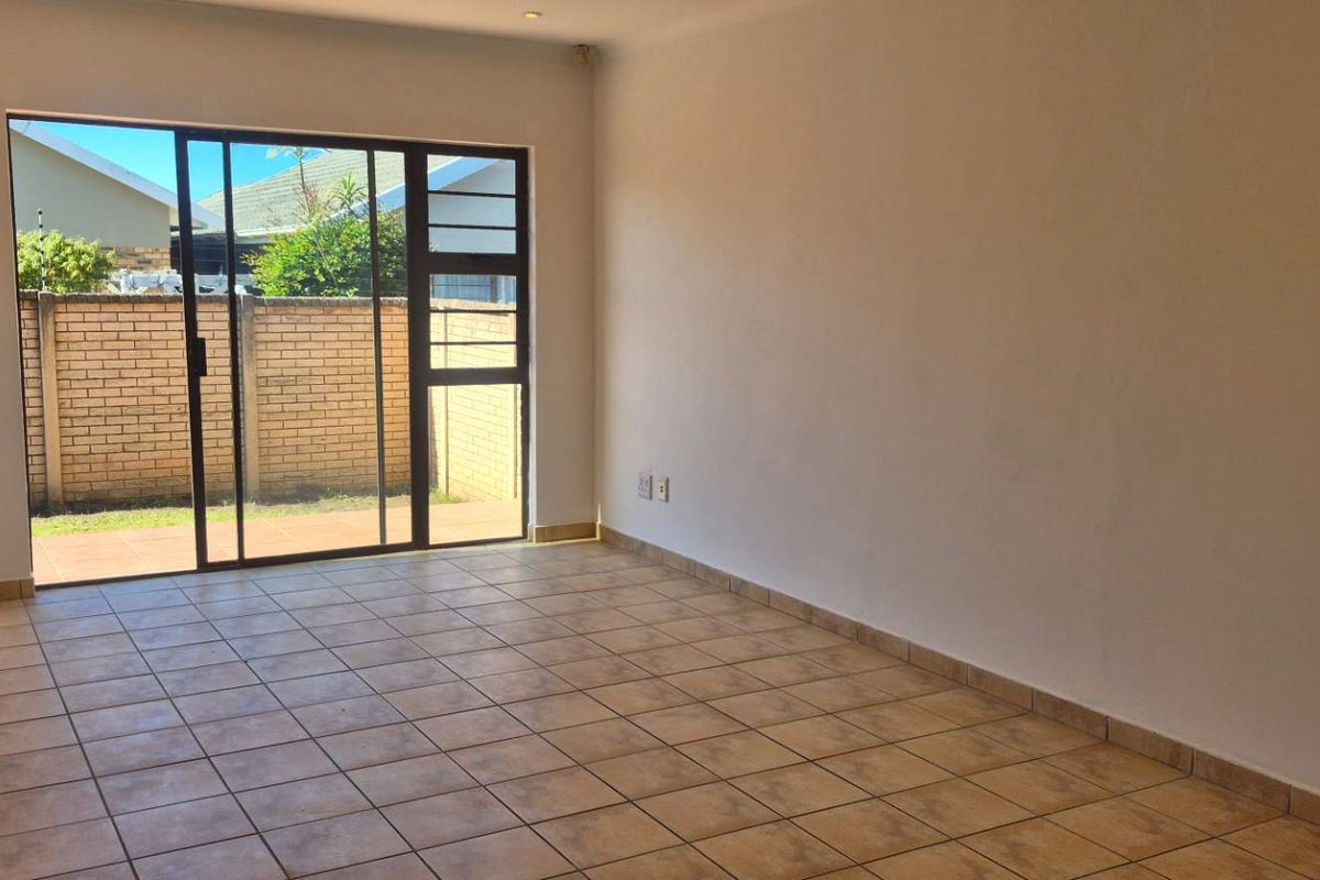 An empty, sunlit living room featuring neutral tiled flooring and a large sliding glass door providing direct access to the outdoor patio area.