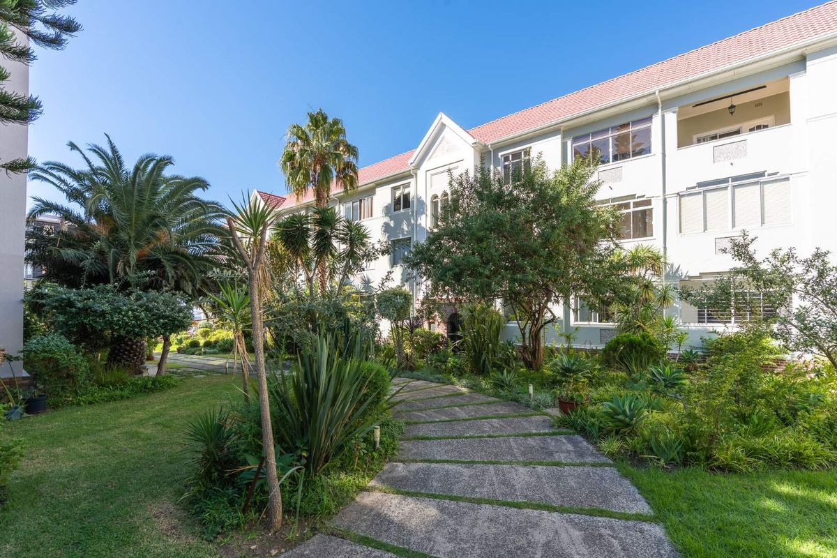 A well-maintained apartment building exterior featuring a lush landscaped garden, paved pathways, and private balconies under a classic tiled roof.