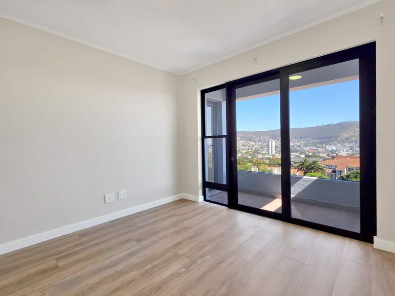 A bright, empty bedroom featuring modern laminate flooring, neutral walls, and a large sliding glass door providing access to a balcony with scenic views.