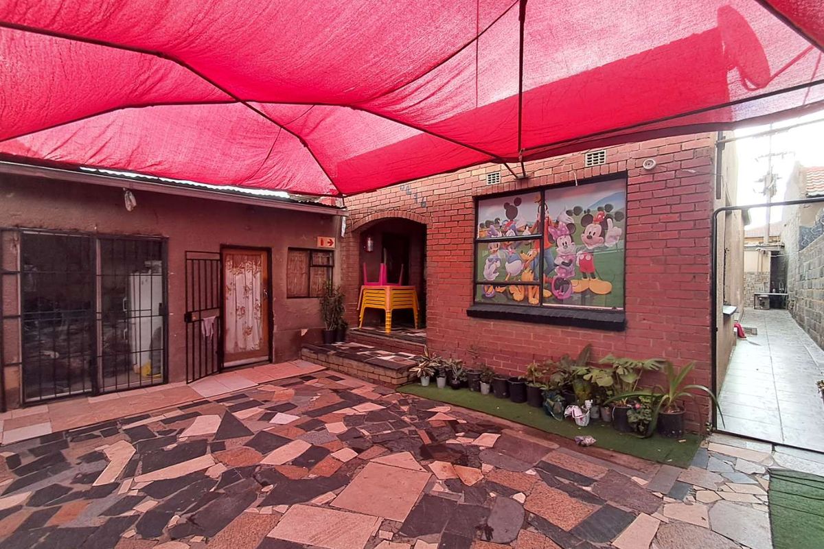 A paved rear courtyard area featuring a large red shade sail, brick facade, and access to the main dwelling.