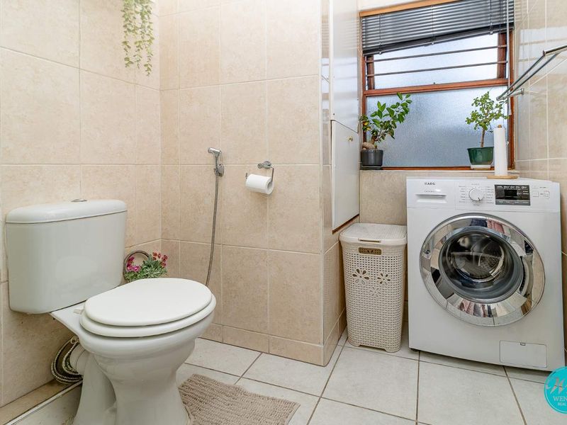 A functional laundry room featuring tiled finishes, a toilet, and space for a washing machine, with a window providing natural light.