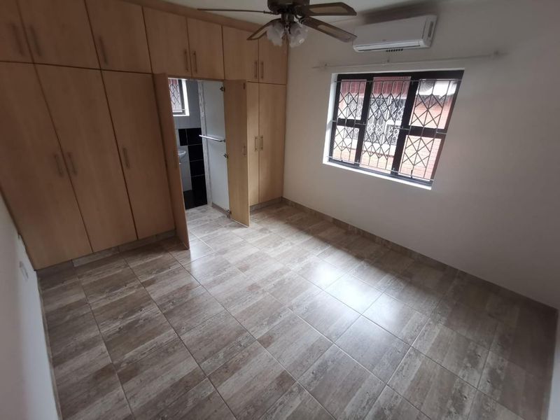 A clean, empty bedroom featuring built-in wooden cupboards, tiled flooring, a ceiling fan, and a wall-mounted air conditioning unit.