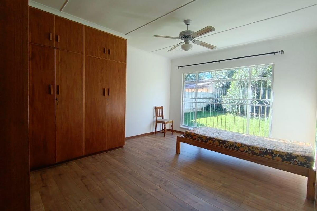 A spacious bedroom featuring built-in wooden cupboards, wood-look flooring, a ceiling fan, and large windows with security bars overlooking the garden.