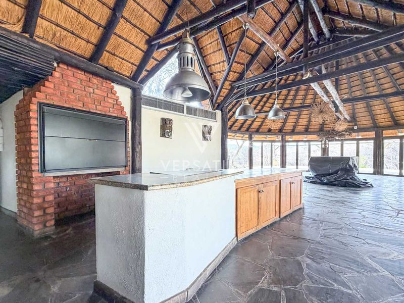 Spacious open-plan entertainment area featuring a high thatched roof with exposed trusses, stone flooring, a built-in brick braai, and a central kitchen island.