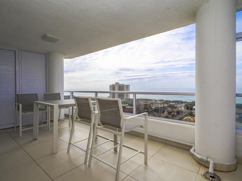 A spacious covered balcony featuring tiled flooring, a glass balustrade, and panoramic coastal views, providing an ideal outdoor living space.