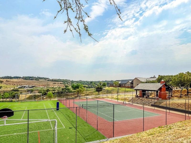 A well-maintained outdoor sports facility featuring a tennis court, soccer field with artificial turf, and a covered pavilion with a brick fireplace.
