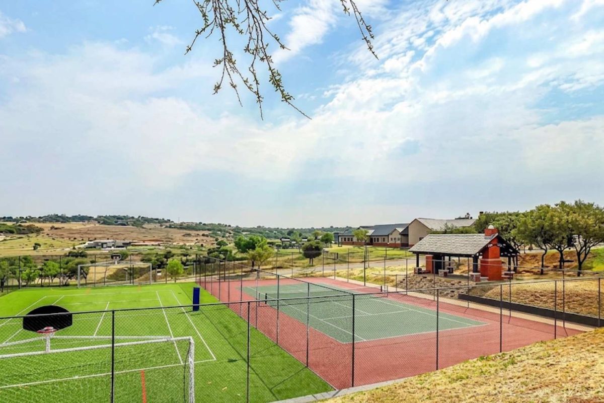 A well-maintained outdoor sports facility featuring a tennis court, soccer field with artificial turf, and a covered pavilion with a brick fireplace.