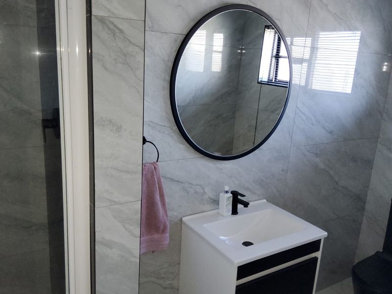 Modern bathroom featuring a vanity cabinet with basin, round mirror, toilet, and a glass-enclosed shower with contemporary grey marble-look wall tiles.