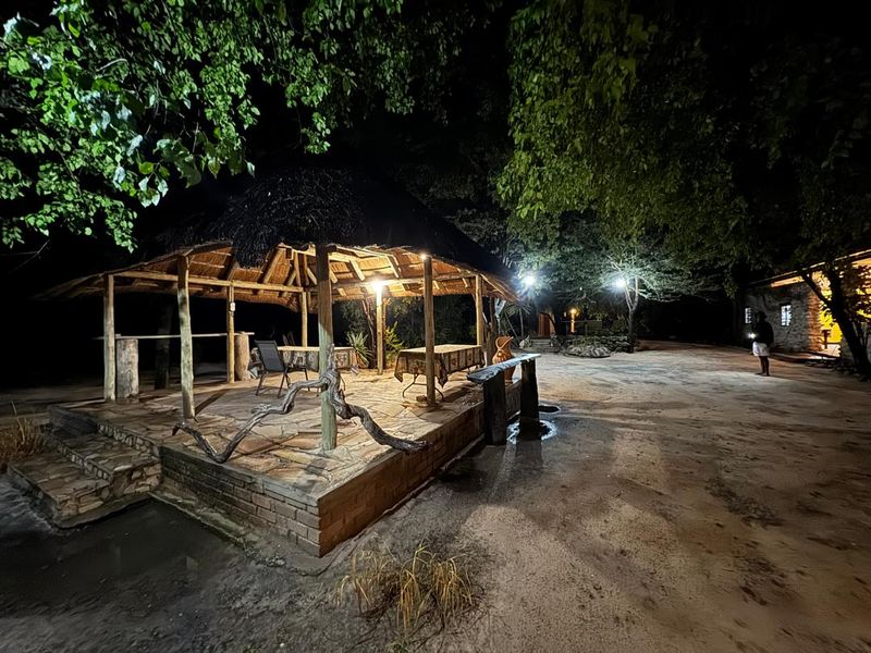 A rustic outdoor entertainment area featuring a thatched roof, stone paving, and timber supports, illuminated for evening use in a farm setting.
