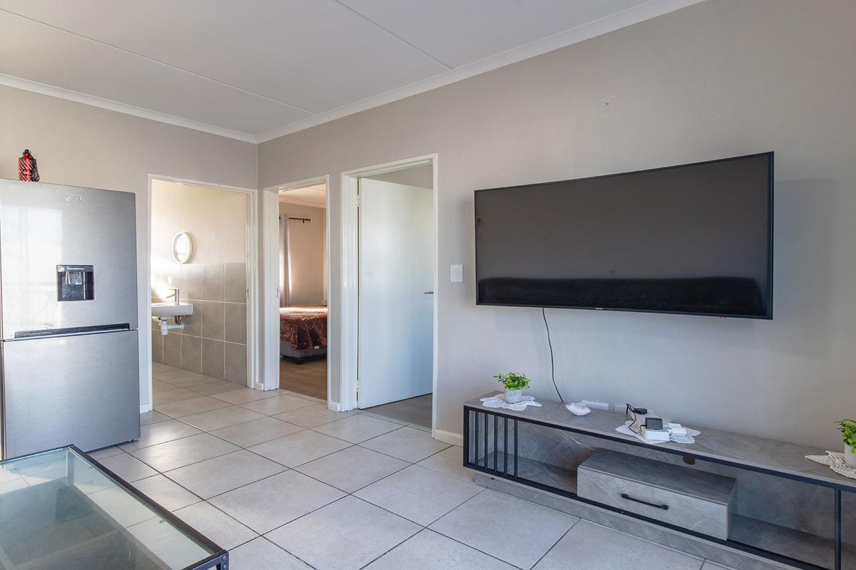 A bright living area featuring neutral tiled flooring, wall-mounted television, and direct access to adjacent rooms.