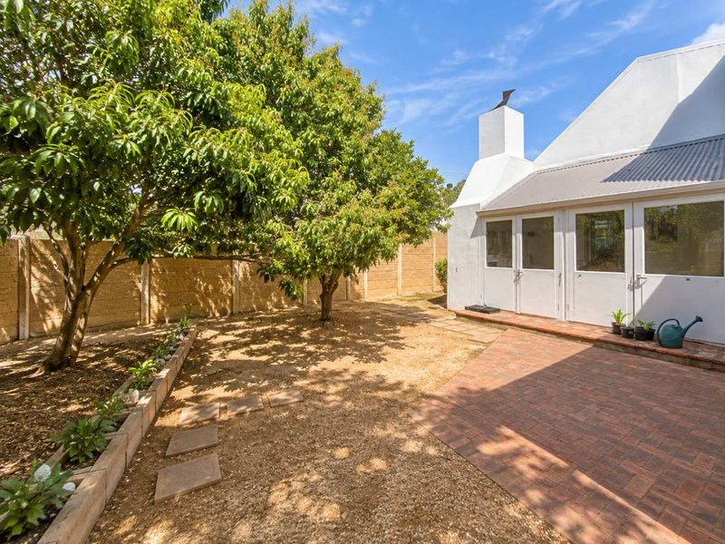 A private rear garden featuring a brick-paved patio area, mature trees, and a white-painted exterior wall with a prominent chimney and glass doors.