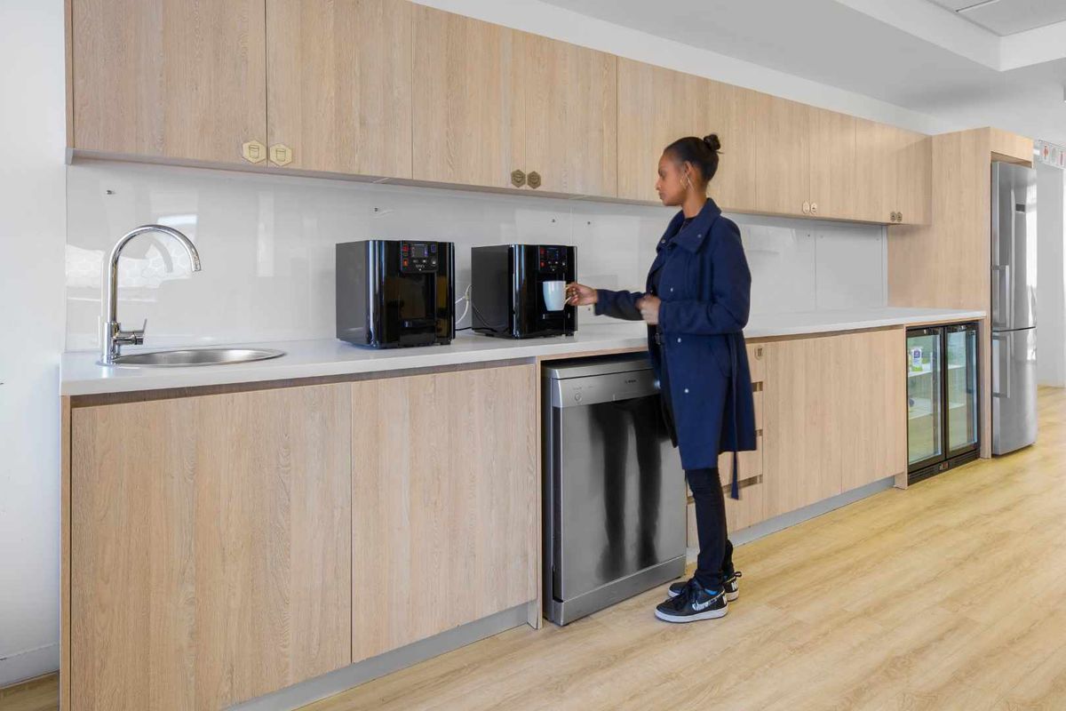 Modern office kitchenette featuring light wood cabinetry, white countertops, a stainless steel sink, and integrated under-counter refrigeration and dishwasher units.