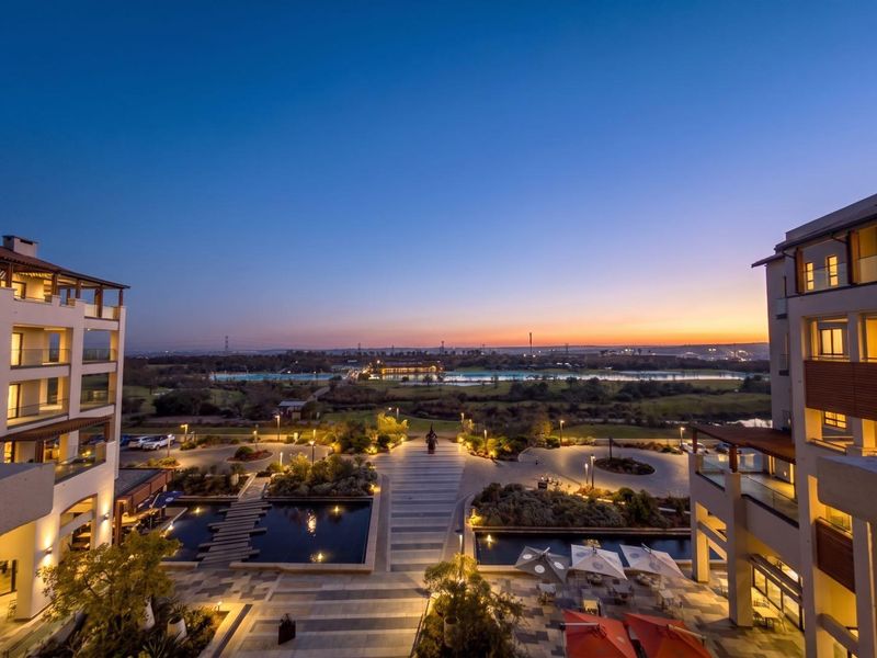A scenic elevated view overlooking the apartment complex grounds, featuring landscaped gardens, water features, and a sunset horizon.