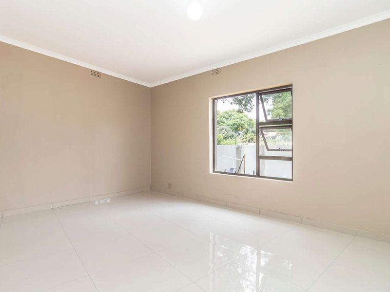 A bright, empty bedroom featuring neutral wall tones, polished floor tiles, and a built-in cupboard with modern aluminium window frames.