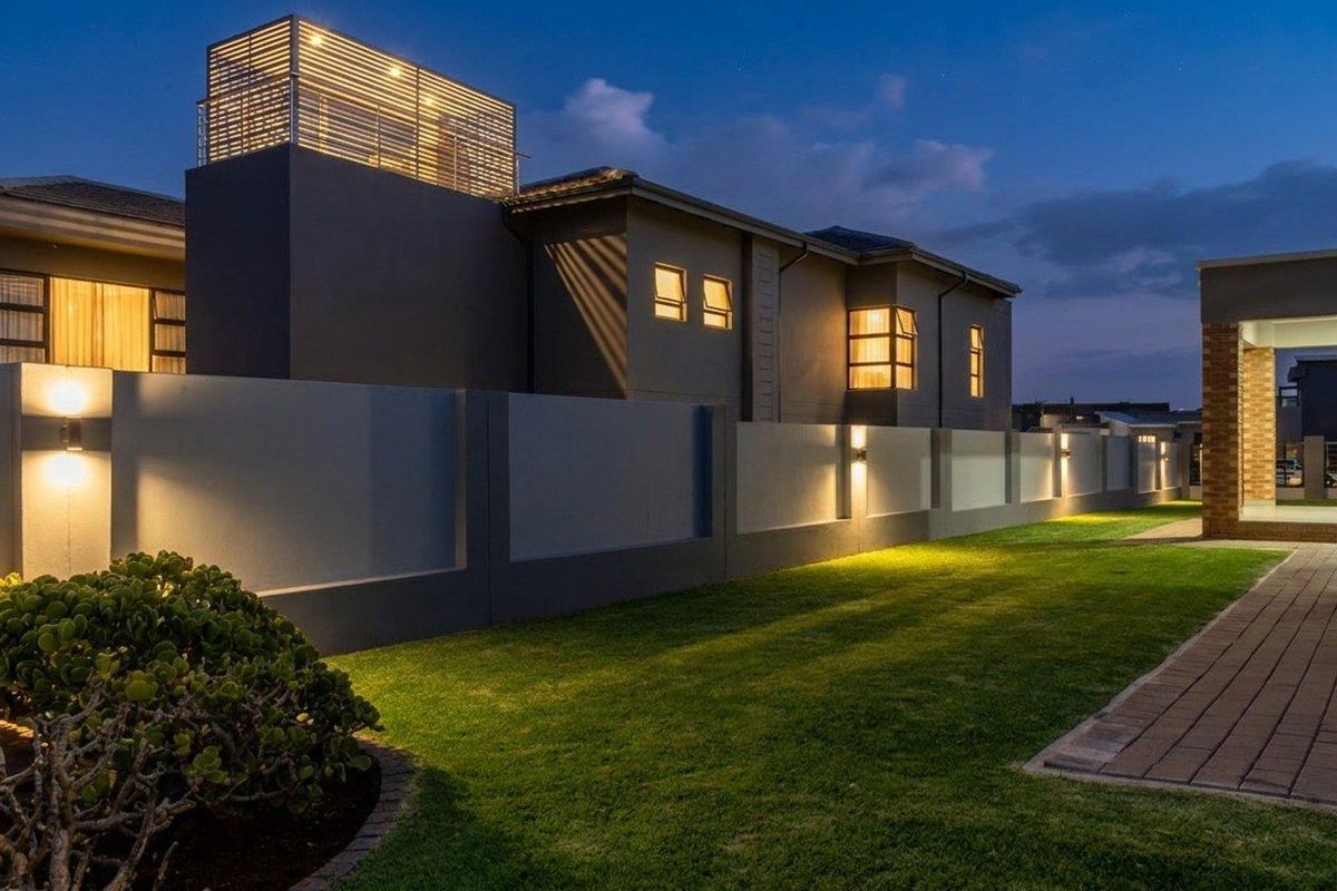 A well-maintained rear exterior view of a modern house at dusk, featuring a paved walkway, landscaped lawn, and integrated wall lighting.