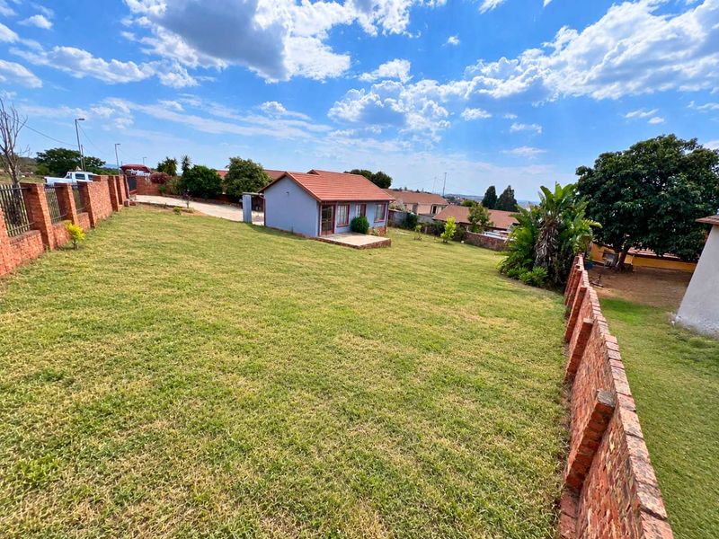 A spacious rear garden featuring a large lawn, brick boundary walls, and a detached dwelling with a tiled roof and small concrete patio.