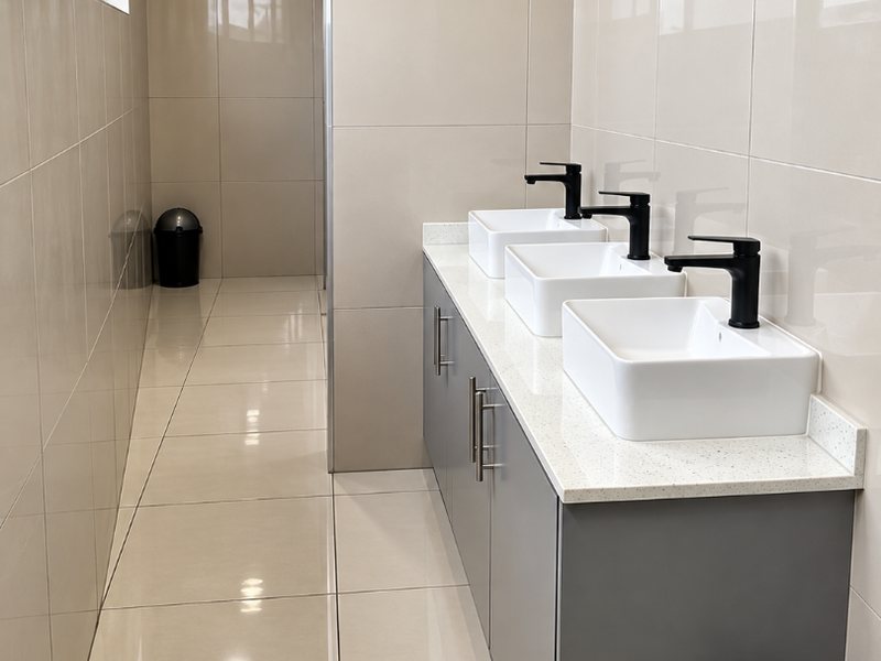 Modern commercial bathroom featuring a triple vanity with vessel sinks, matte black fixtures, and neutral floor-to-ceiling tiling.