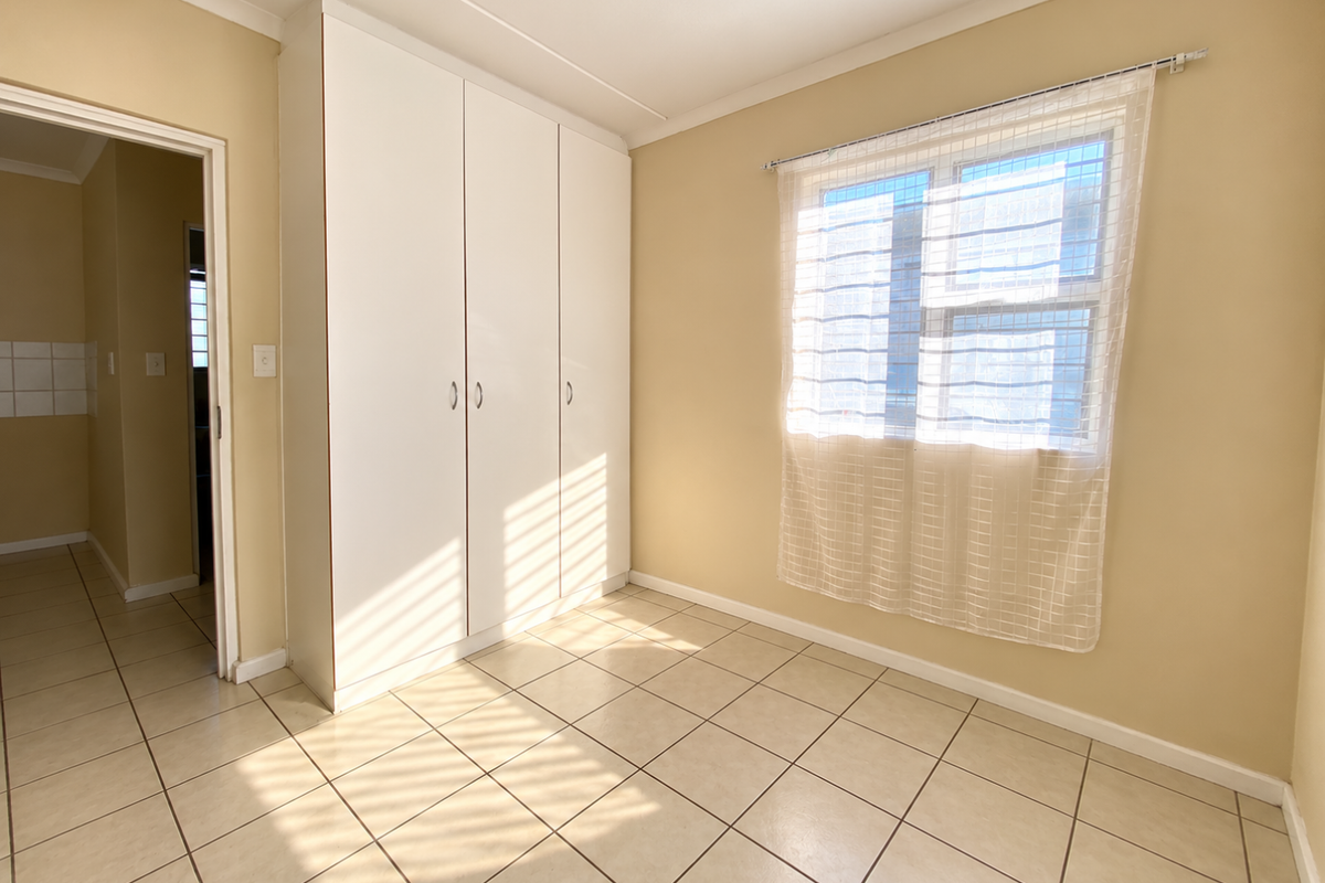 A bright, empty bedroom featuring neutral tiled flooring, a built-in white wardrobe, and a window with natural light.