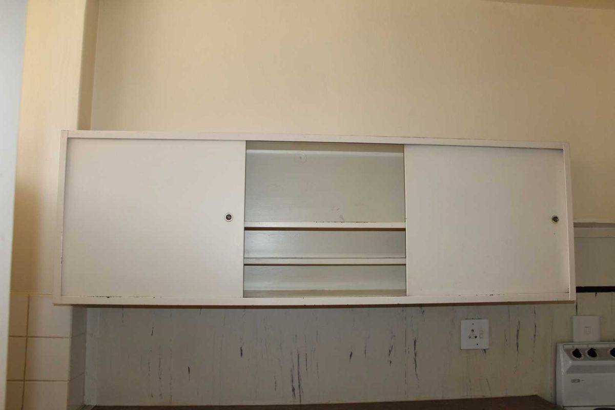 A dated kitchen wall cabinet with sliding doors, featuring a tiled splashback and visible wear on the surrounding wall surfaces.