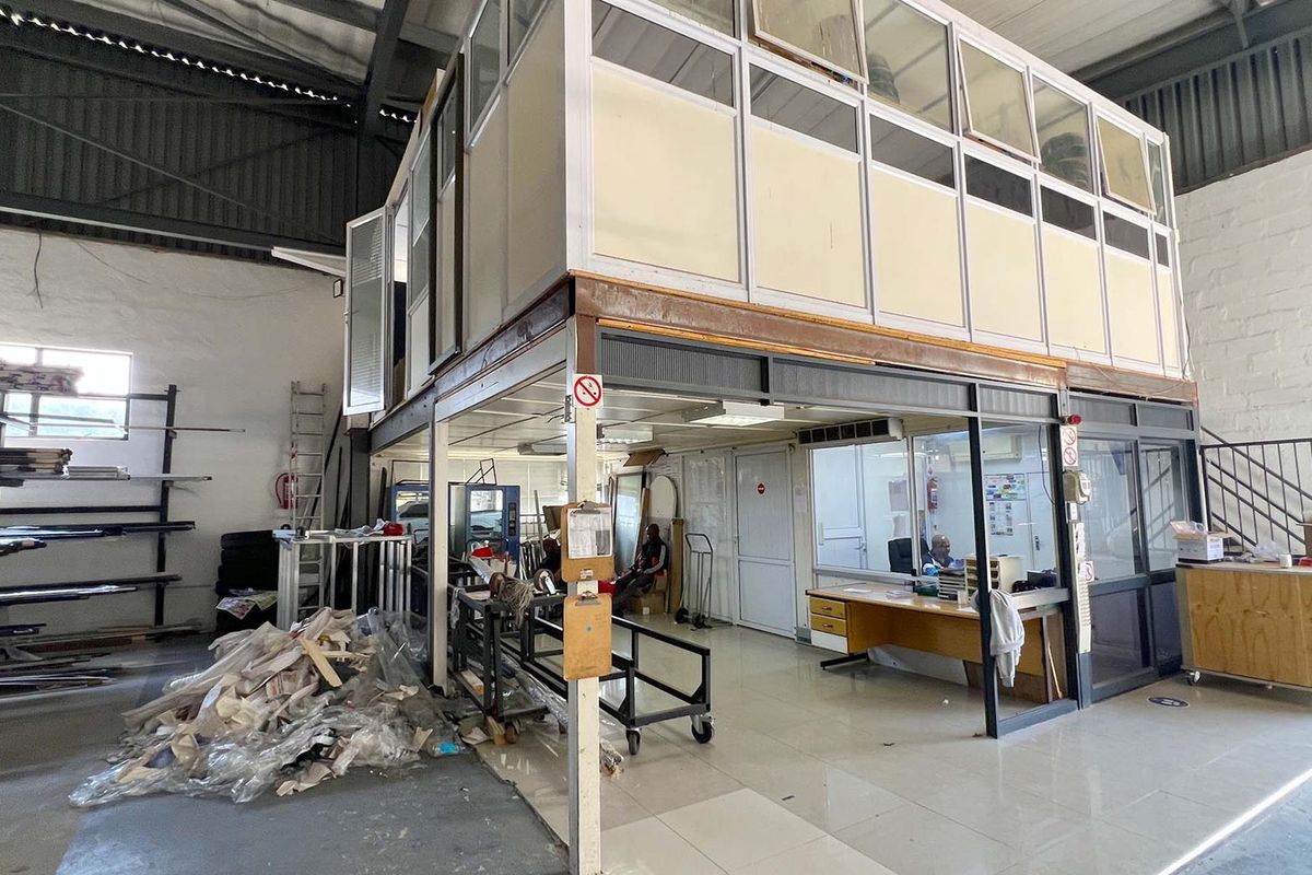 Industrial warehouse space featuring a built-in mezzanine office structure with partitioned glazing, tiled flooring, and high-clearance exposed steel roof trusses.