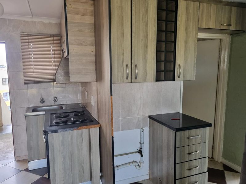 A compact kitchen featuring wood-look cabinetry, an electric hob, and tiled backsplash, currently requiring minor repairs to cabinetry and exposed plumbing.