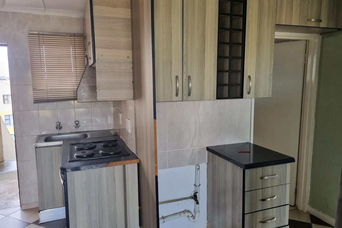 A compact kitchen featuring wood-look cabinetry, an electric hob, and tiled backsplash, currently requiring minor repairs to cabinetry and exposed plumbing.
