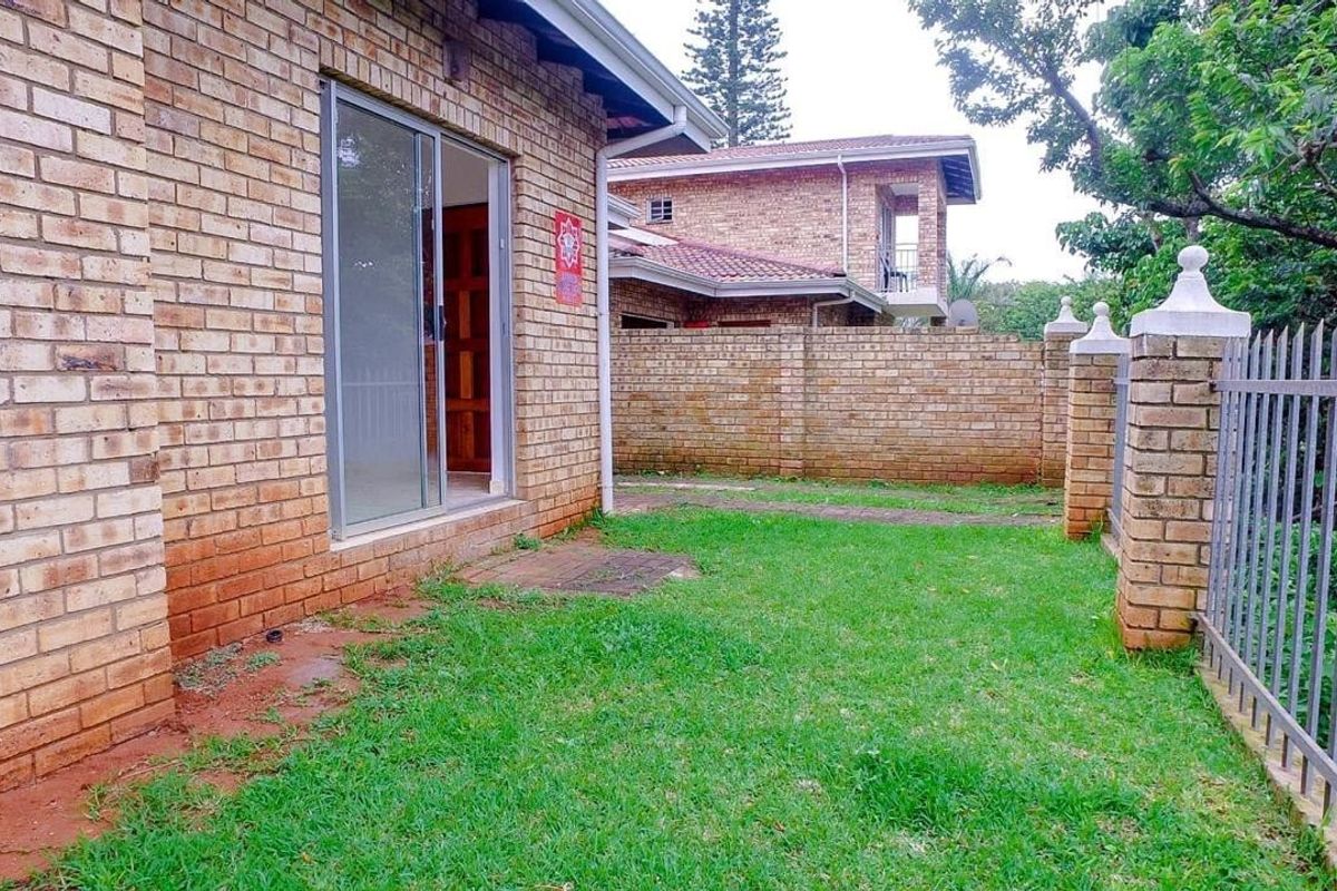A rear exterior view of a face brick townhouse featuring a sliding glass door, a private lawn area, and secure brick and metal perimeter fencing.