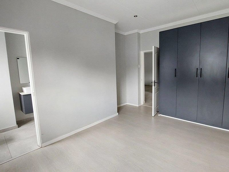 A bright, empty bedroom featuring modern dark grey built-in cupboards, light laminate flooring, and an en-suite bathroom.