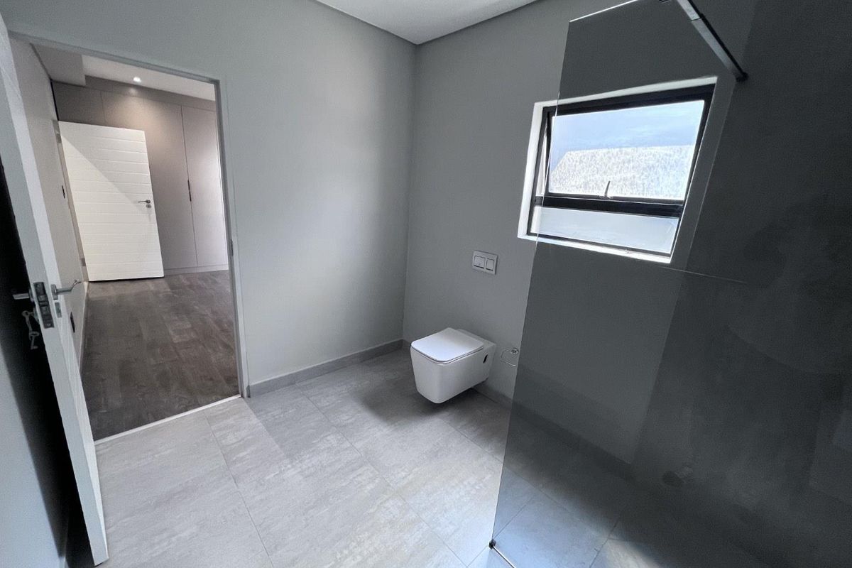 Modern bathroom featuring a wall-mounted toilet, glass shower screen, and neutral floor tiling, with direct access to an adjacent bedroom.