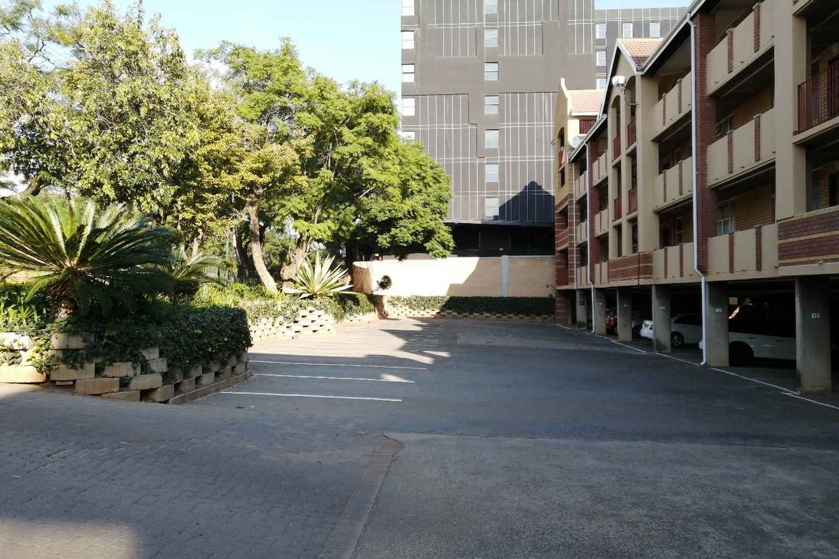 Exterior view of the apartment complex showing a paved parking area, covered parking bays, and landscaped garden beds.