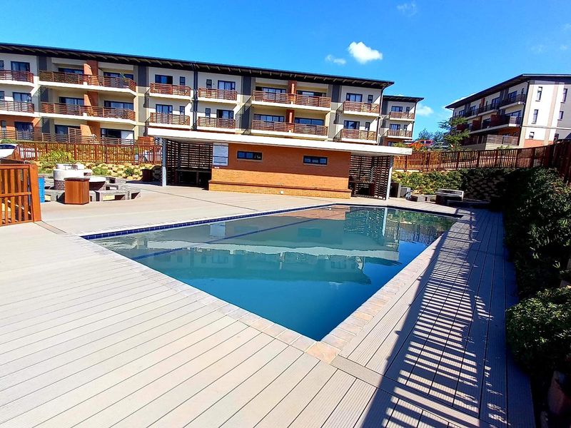 A well-maintained communal swimming pool area featuring composite decking, brick clubhouse facilities, and secure timber fencing within an apartment complex.
