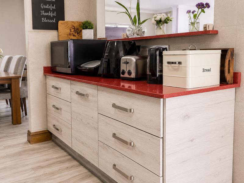 A modern kitchen area featuring light wood grain cabinetry, a vibrant red quartz countertop, and a neutral tiled splashback.