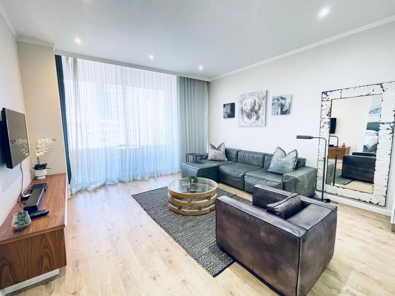 A well-maintained living room featuring light-toned laminate flooring, recessed downlighting, and floor-to-ceiling sheer curtains providing natural light.