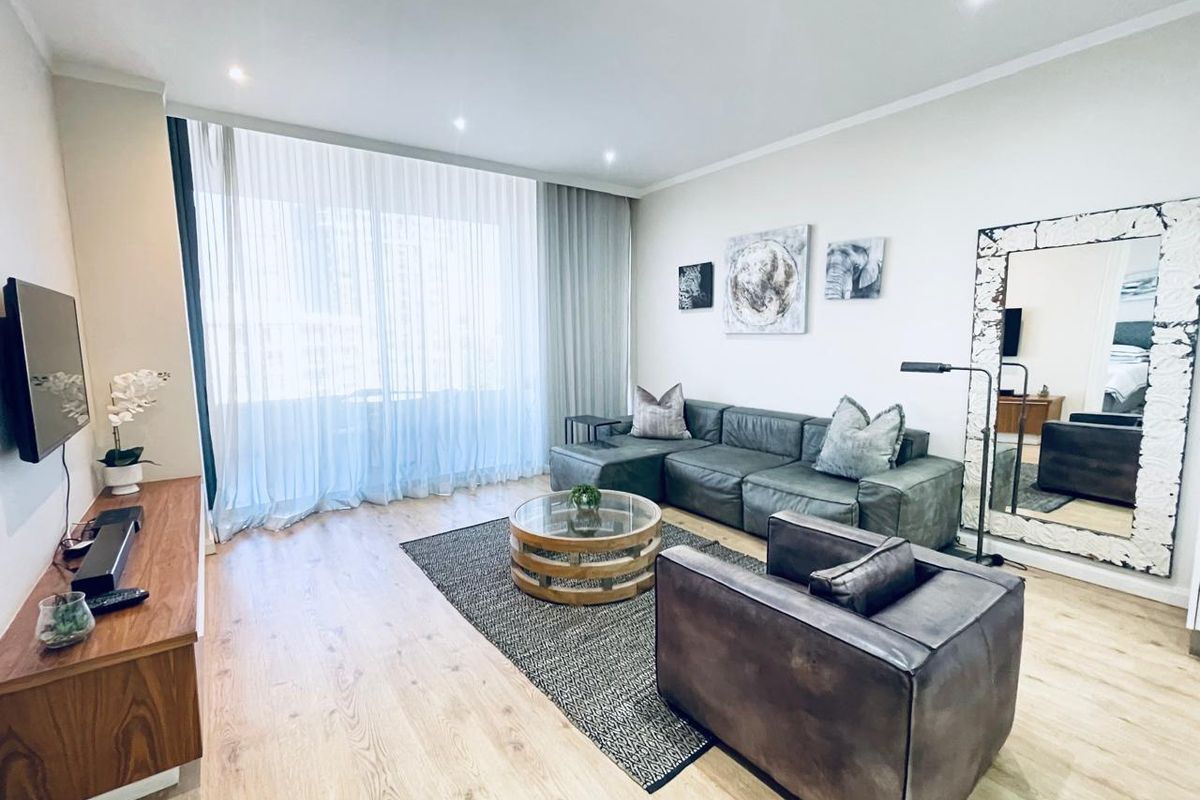 A well-maintained living room featuring light-toned laminate flooring, recessed downlighting, and floor-to-ceiling sheer curtains providing natural light.