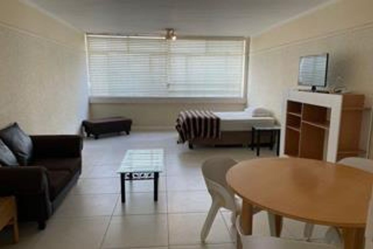 An open-plan living and sleeping area featuring tiled flooring, large windows with venetian blinds, and a functional layout suitable for versatile use.
