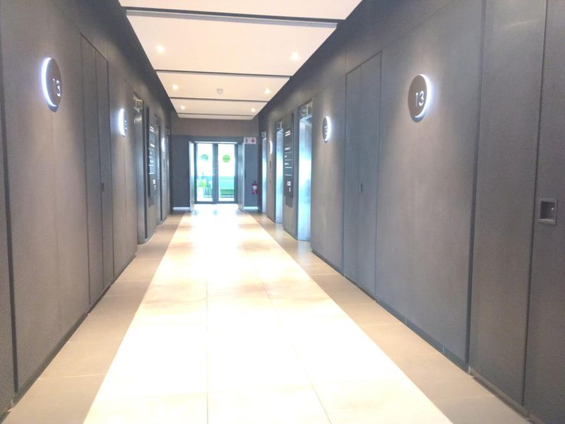 A modern, well-lit office building hallway featuring tiled floors, recessed ceiling lights, and multiple elevator access points with sleek wall finishes.