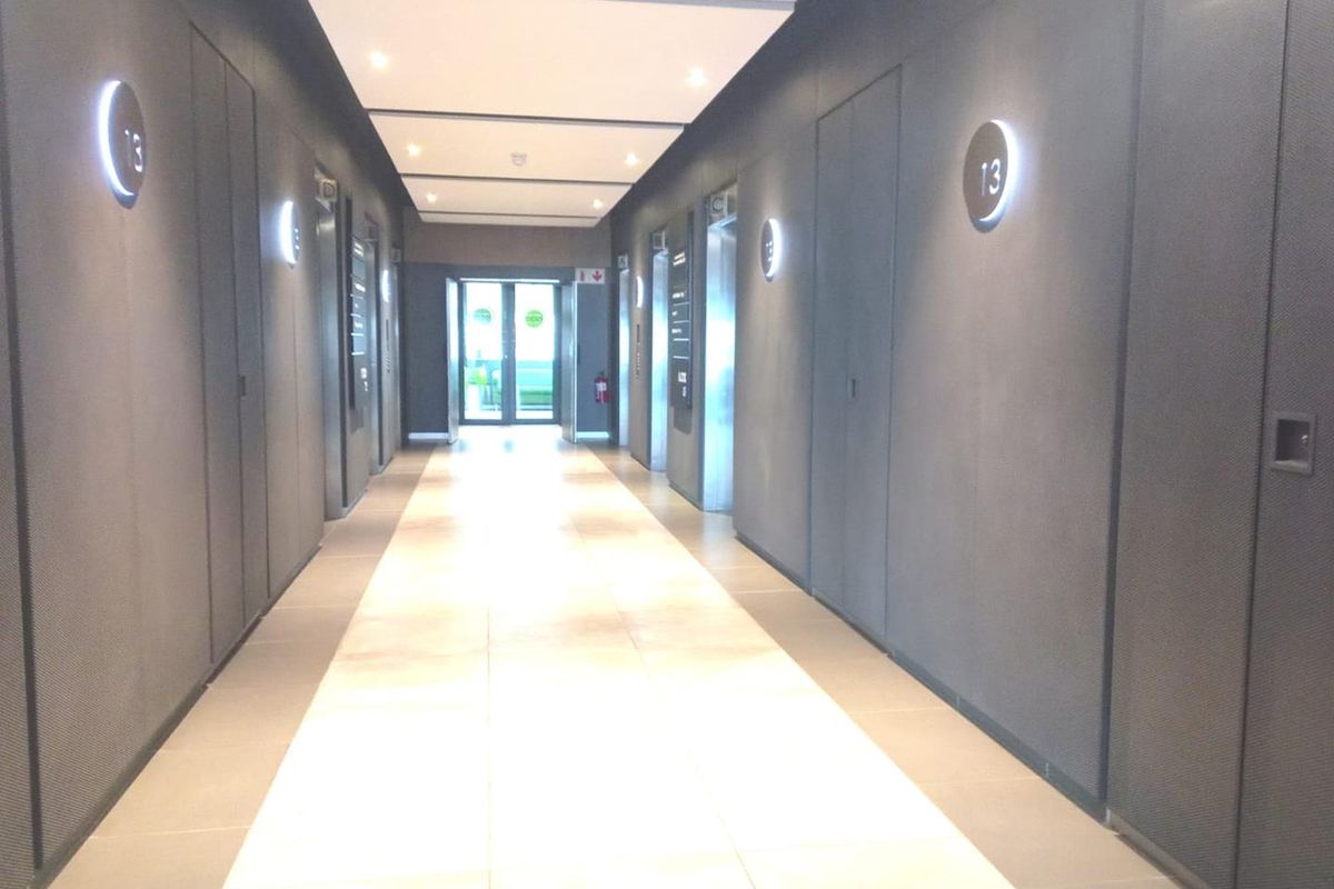 A modern, well-lit office building hallway featuring tiled floors, recessed ceiling lights, and multiple elevator access points with sleek wall finishes.